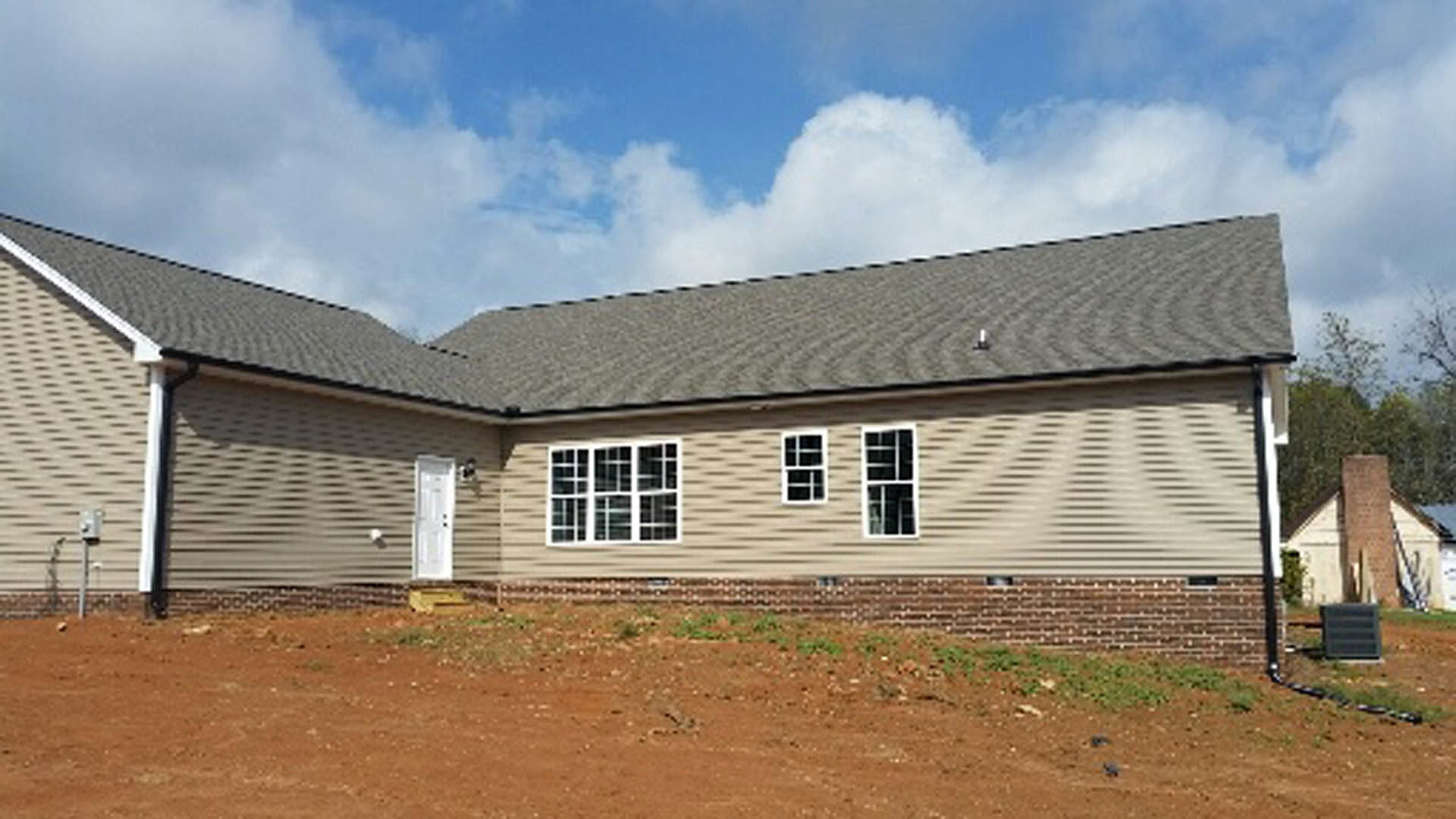Single-story home with light siding, white door, and multiple windows, set behind a fenced dirt field under a partly cloudy blue sky