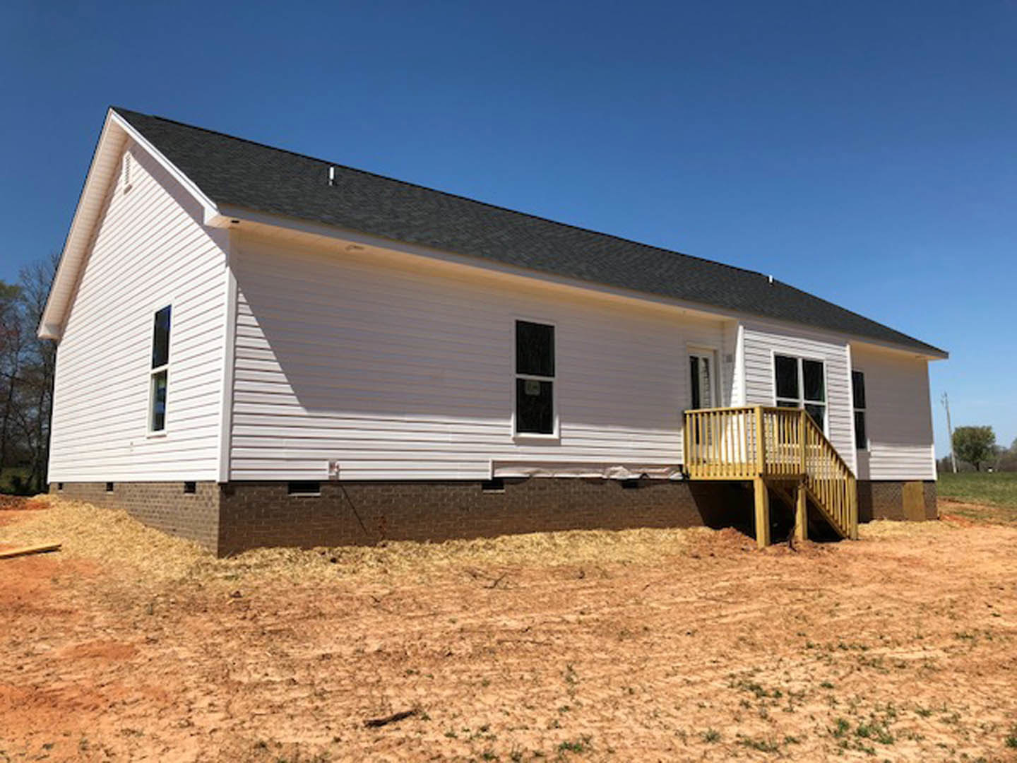 White siding house with yellow railing porch, wooden deck and staircase, dirt field with sparse grass in front, multiple windows visible.