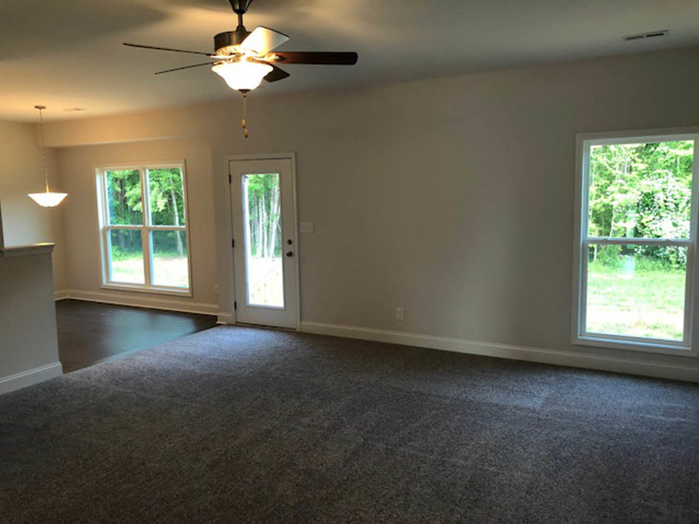 Neutral-toned room featuring a ceiling fan with light fixture, multiple windows showing leafy trees outside, white door with glass panel, and smooth flooring.