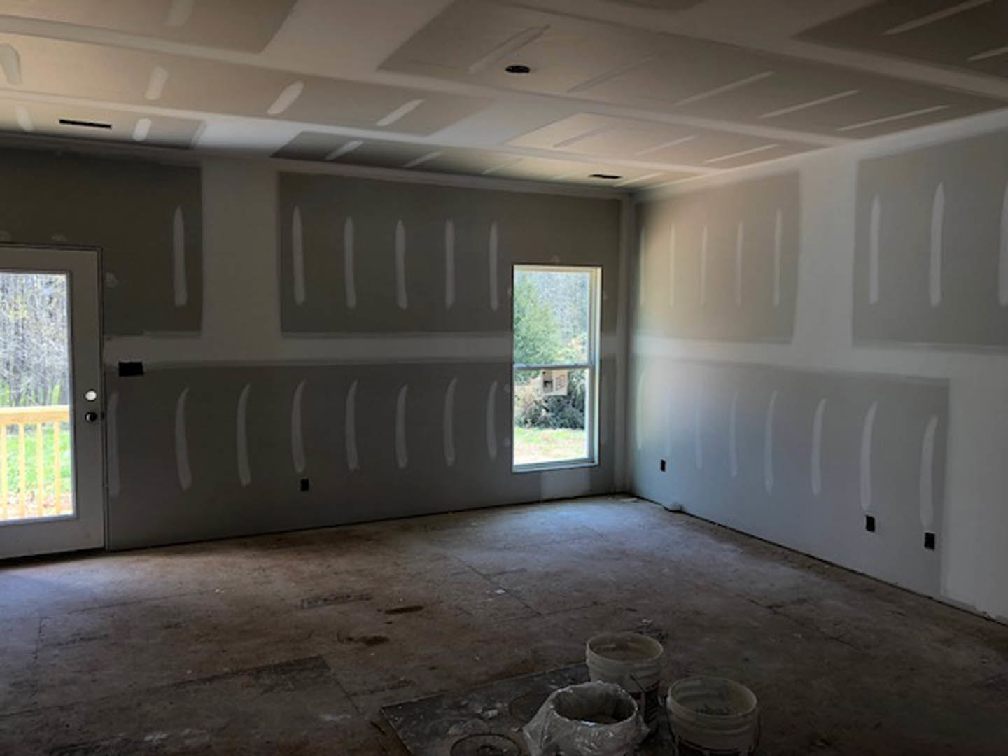 White-painted walls and ceiling, large window with tree views, unfinished floor with buckets, plaster finishes, indoor residential space.