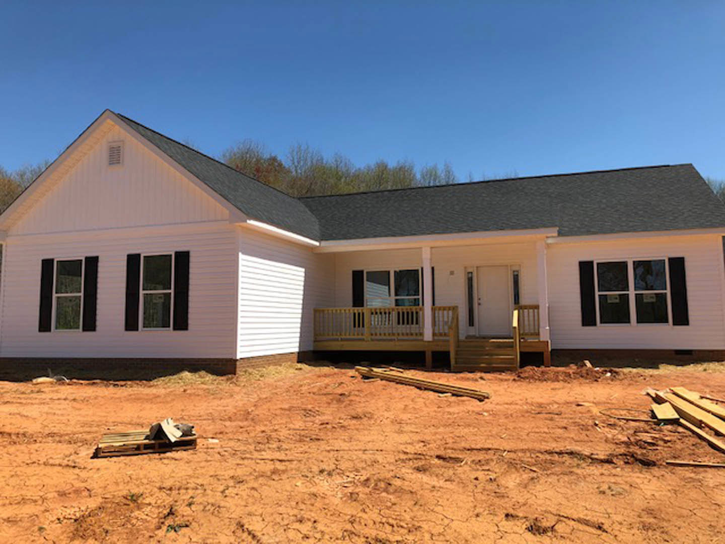 White house under construction with dirt ground, pile of wood, white porch railing, glass-paneled white door, and white-framed window under blue sky