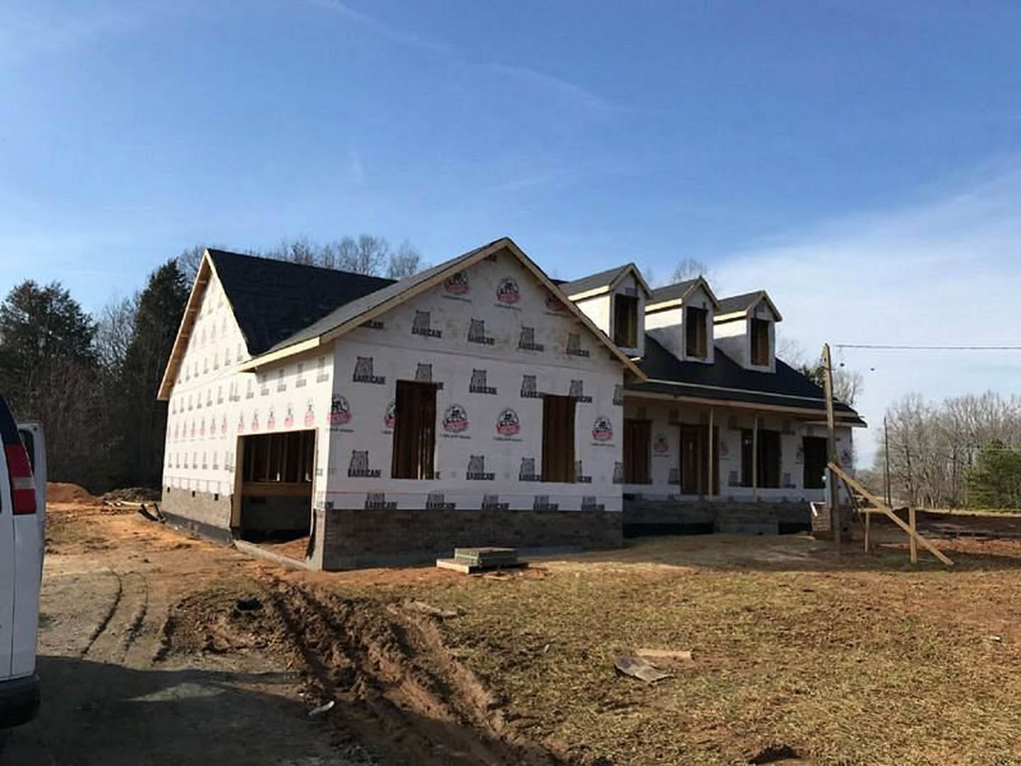 Partially built house with white exterior walls covered in black construction logos, dirt path leading to entrance, wooden fence in yard, scattered construction materials, cloudy
