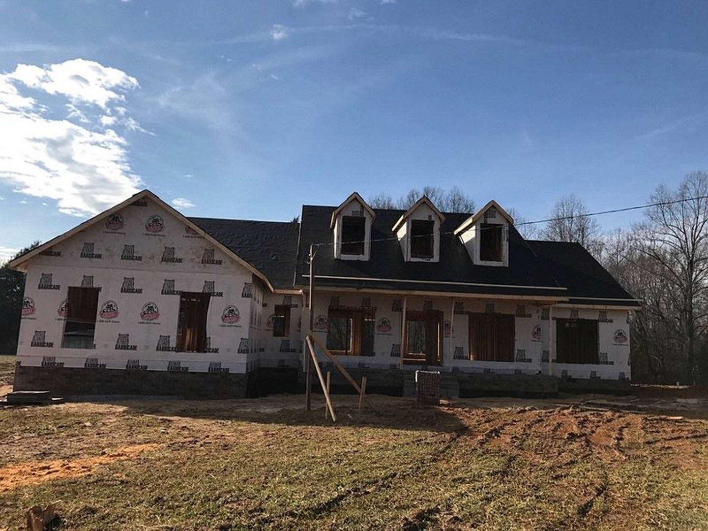 Modern home under construction with black roof, exposed wooden framing, large windows, grassy yard, and blue sky with scattered clouds