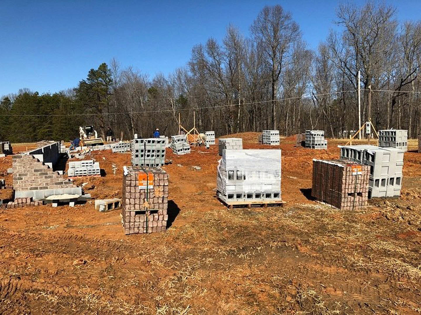 Stack of brown bricks on a grassy field with leafless trees and a white tractor in the background, under an open sky.