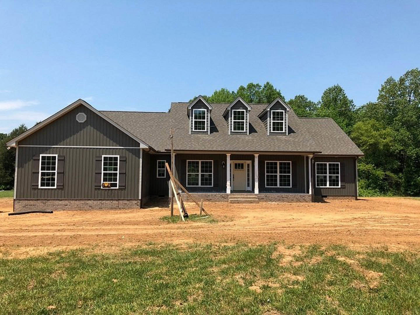 Grey custom home with white-framed windows, unfinished dirt yard, ladder leaning against exterior, and partial view of door; sparse grass and trees in background.