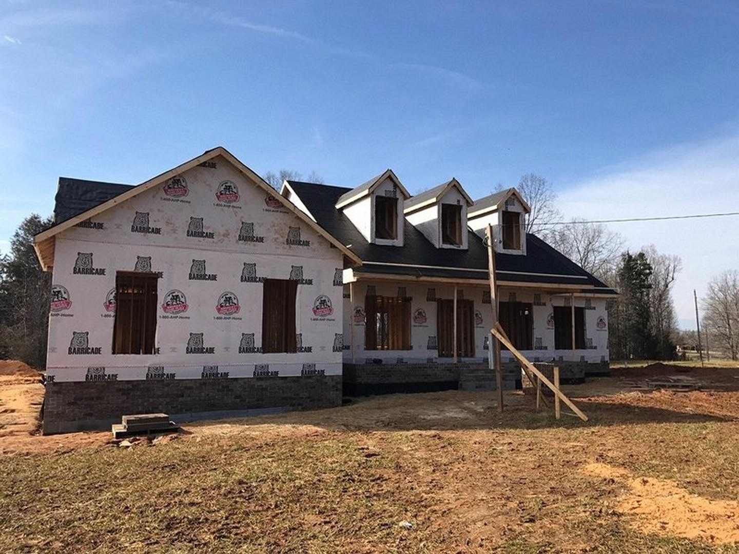 Modern home under construction with white exterior walls featuring black and red logos, broken wooden post and temporary fencing in foreground, grassy yard, large windows, and