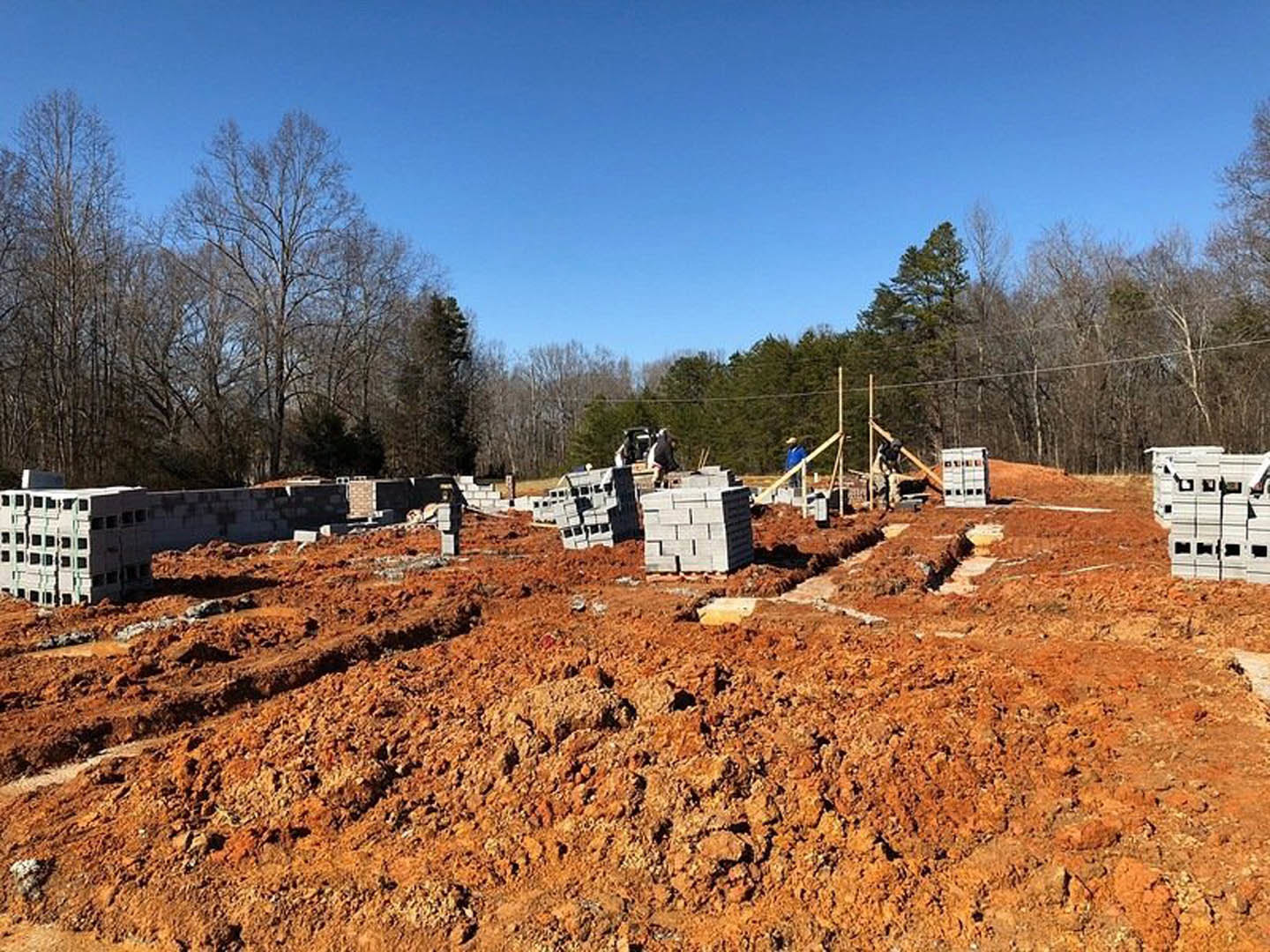 Bricks stacked on dirt at a residential construction site, partially built brick wall, white plastic crates, trees and blue sky in background