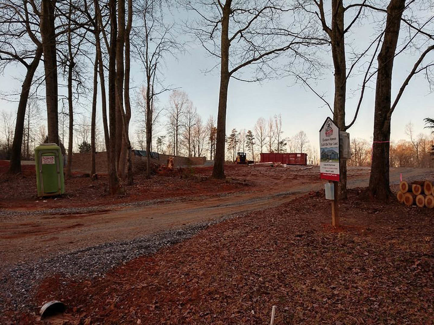 Dirt road lined with leafless deciduous trees, green portable toilet with attached sign, stack of cut logs, distant building partially visible, tractor parked among trees in winter