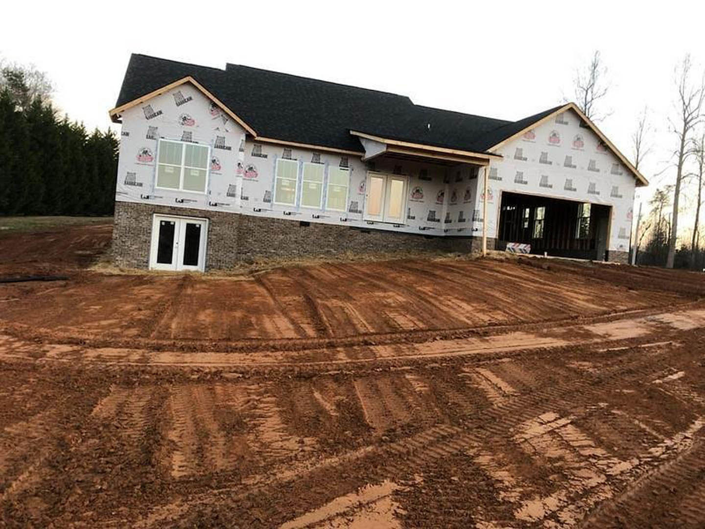 Partially built home with black roof, white double doors featuring black glass panels, surrounded by dirt lot and mature trees, tire tracks visible on ground