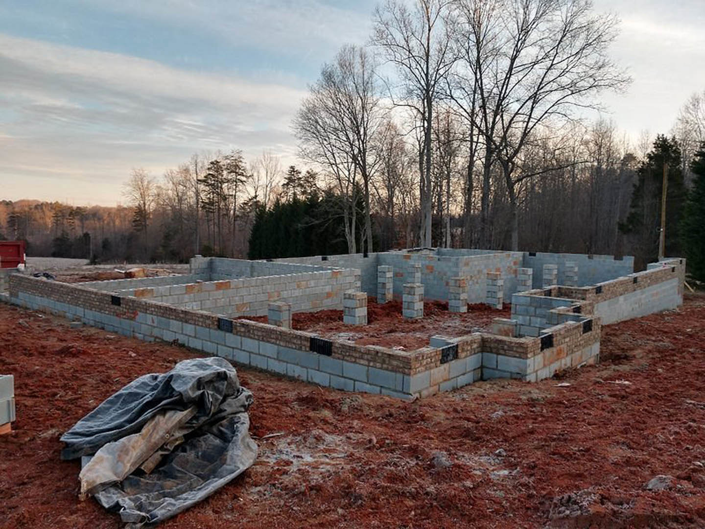Construction site with partially built brick walls, stacked bricks on bare ground, blue tarp covering materials, and trees in the background under a cloudy sky.
