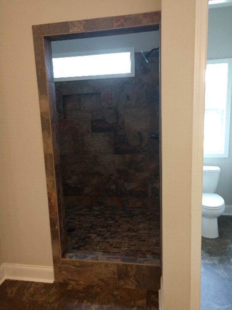Walk-in shower with gray tile floor, glass enclosure, white tile walls, and chrome fixtures adjacent to a white toilet and blue-trimmed door