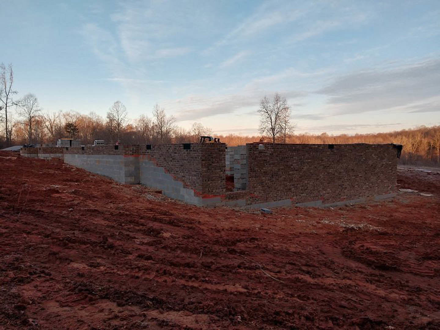 Brick home under construction surrounded by red dirt, leafless trees, and a partly cloudy blue sky