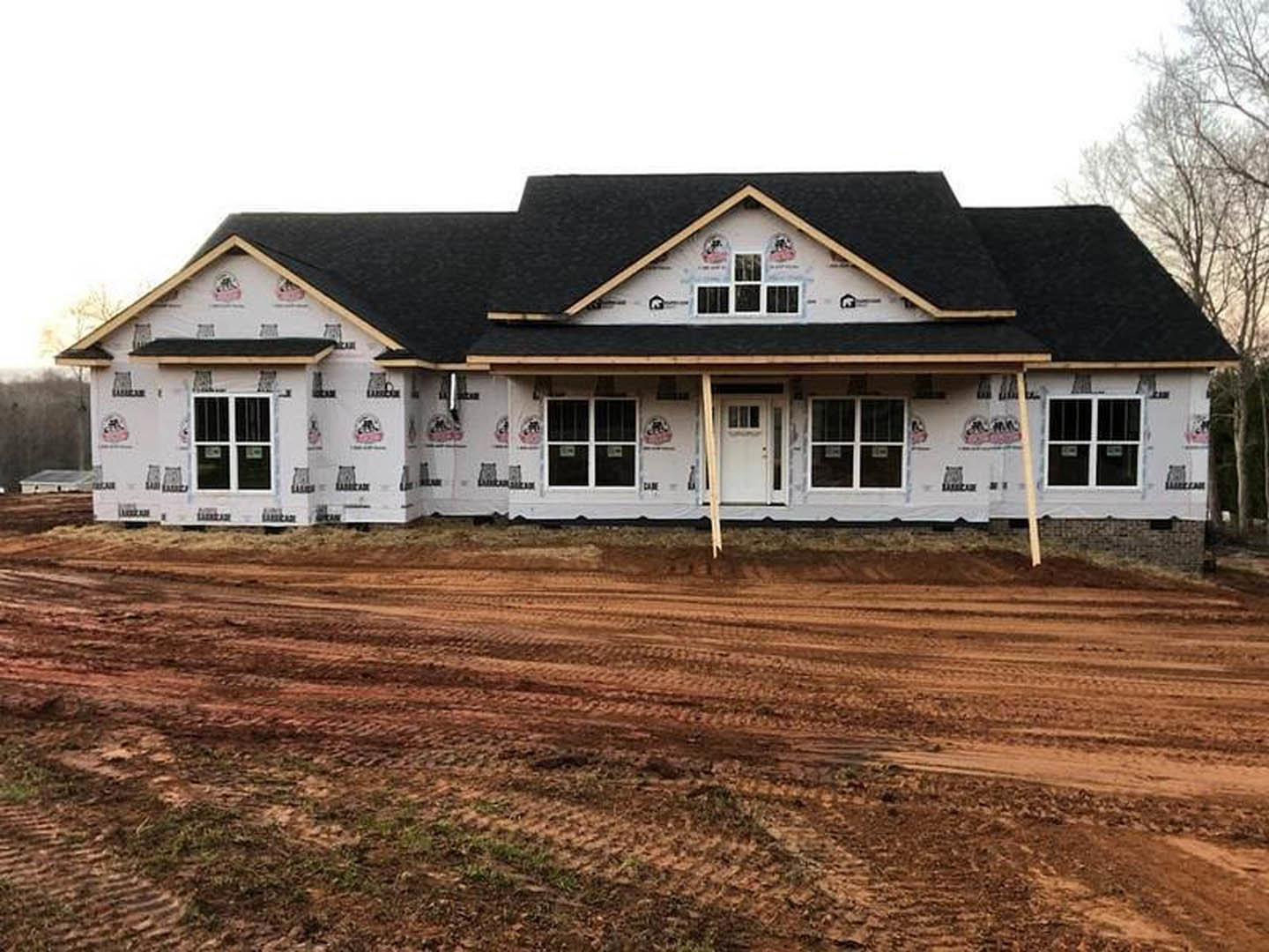 Two-story house under construction with black roof, white siding, and large windows, surrounded by dirt and mud with visible tire tracks