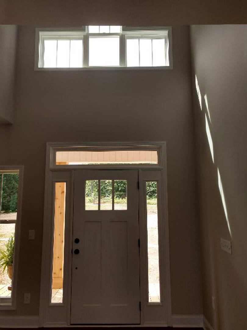 White paneled door with glass inserts, adjacent window letting in natural light, white wall outlet below, view of green trees through window, smooth white walls.