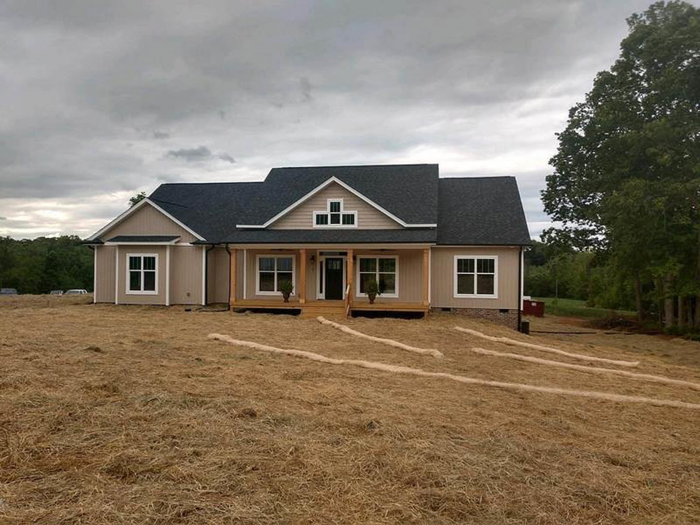 Partially built house with black roof and few windows, surrounded by expansive grassy field and leafy tree under cloudy sky