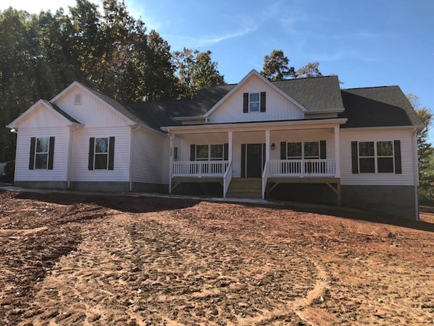 Two-story house with white-framed windows, covered porch with white railing, large grassy yard bordered by trees, blue sky overhead