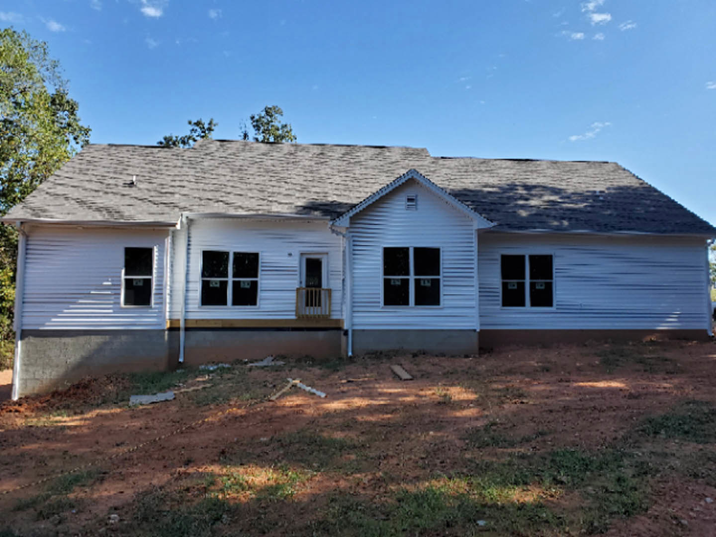 Two-story house under construction with exposed framing, dirt yard, covered porch, and blue sky overhead