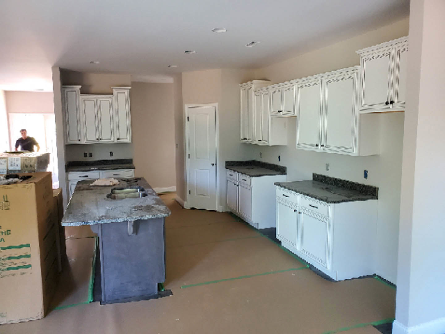 White kitchen with white cabinets and countertops, black hardware on a white door, cutout in the countertop for sink installation, blurred figure in background, white box with