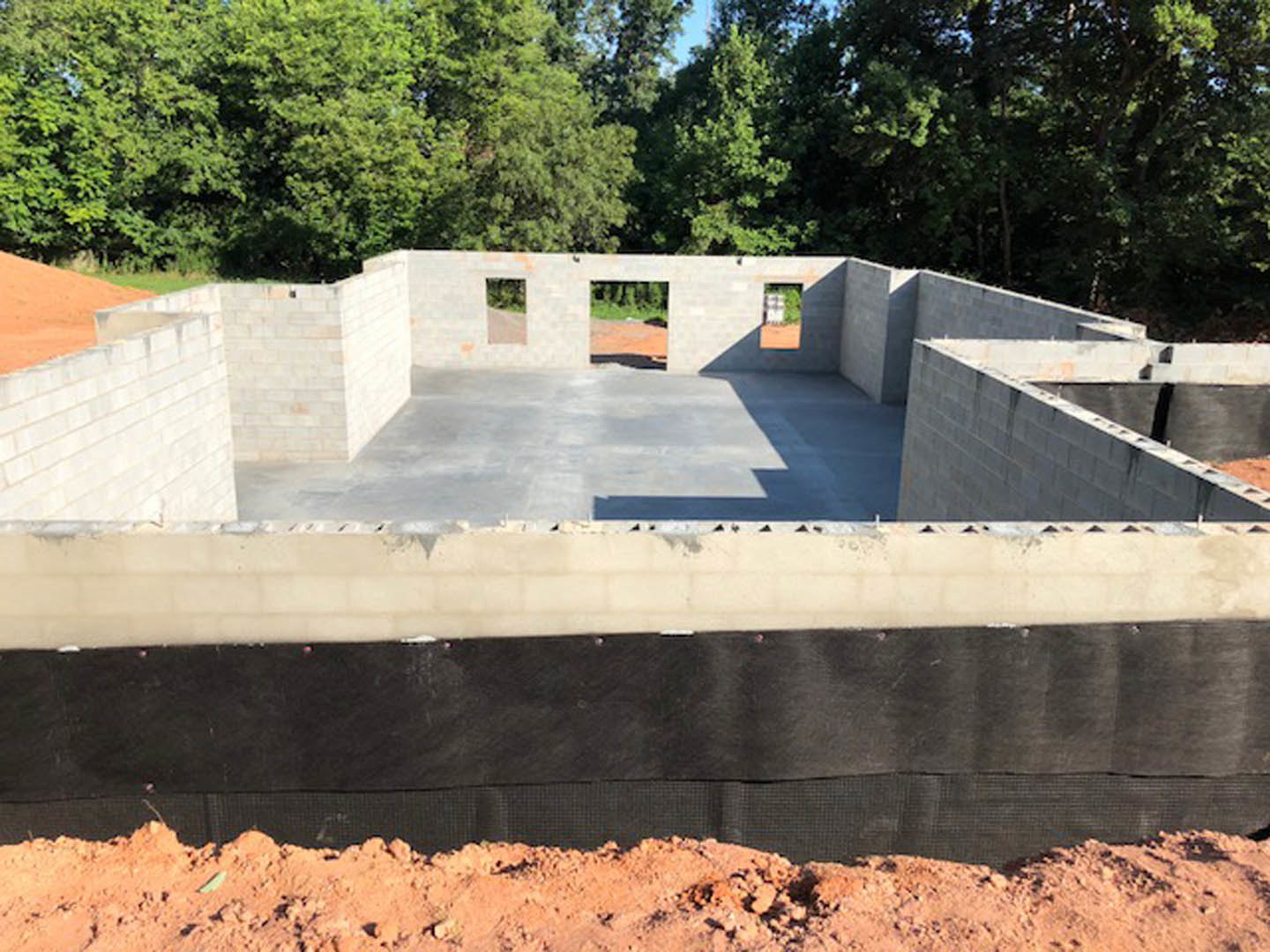 Concrete foundation with white wall and rectangular windows, surrounded by dirt ground and trees in the background