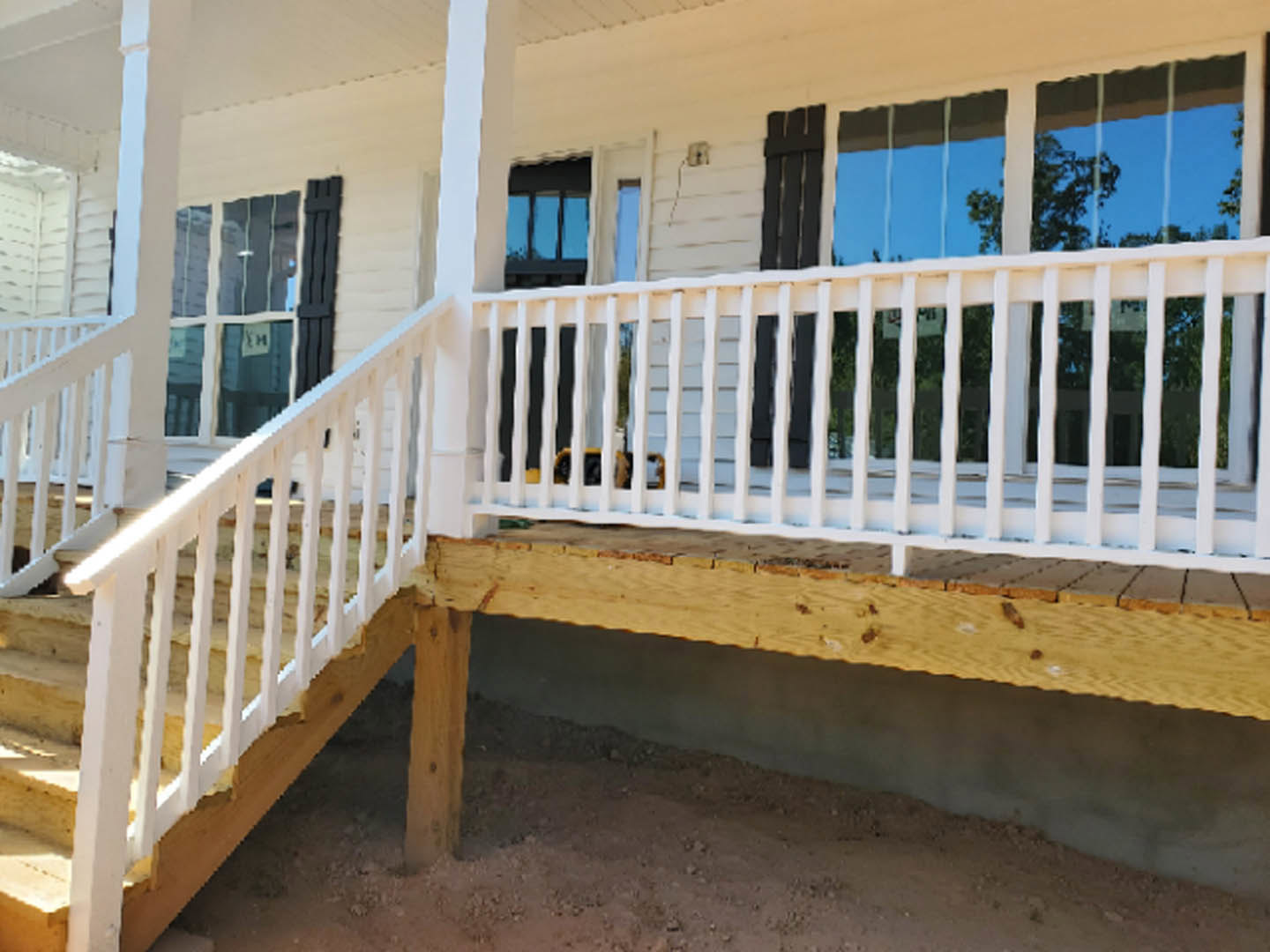 White railing with square balusters on a wooden porch, concrete foundation below, window reflecting blue sky, white pillar partially visible