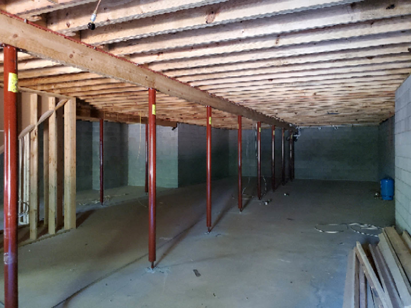 Wooden ceiling with exposed beams, red steel support poles, concrete floor, unfinished walls, and stacked wood planks in a basement interior.