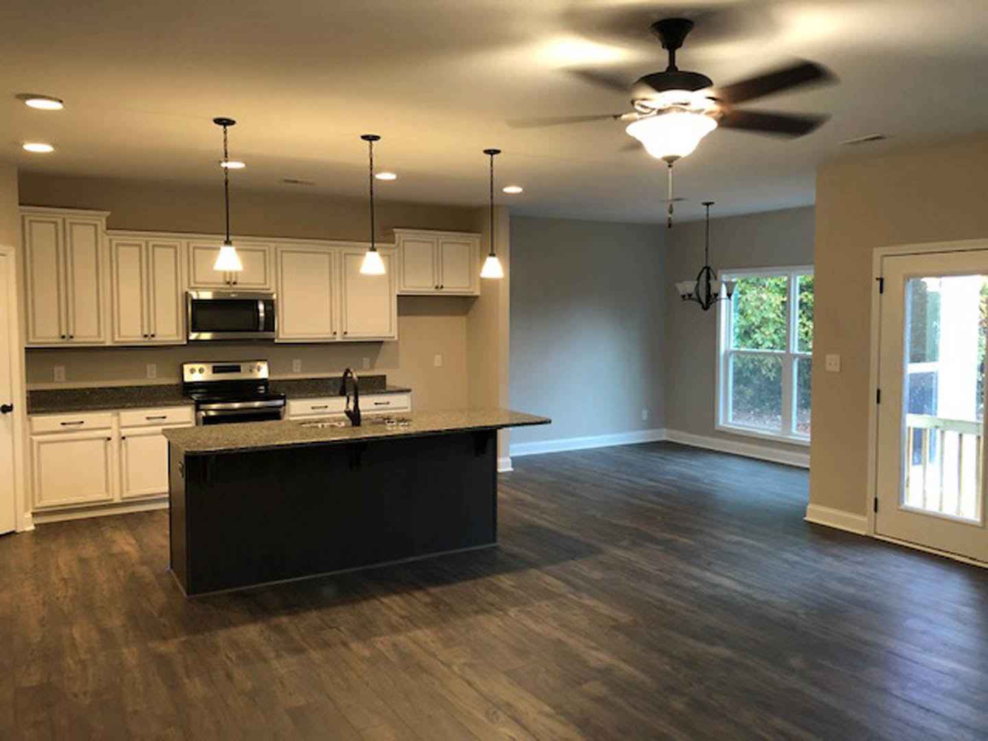 Open kitchen and dining area featuring wood flooring, black countertop, white cabinetry, stainless steel microwave and oven, ceiling fan, and a window with a white frame.