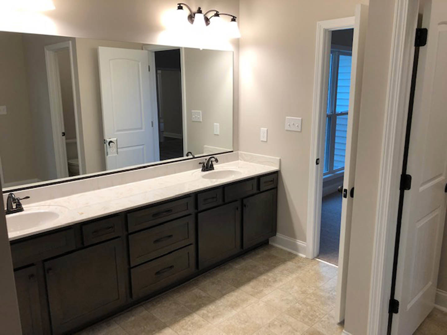 Bathroom with expansive mirror above double sink vanity, white tile backsplash, modern faucets, light cabinetry, and partial view of white bathtub and triple-knob light switch.