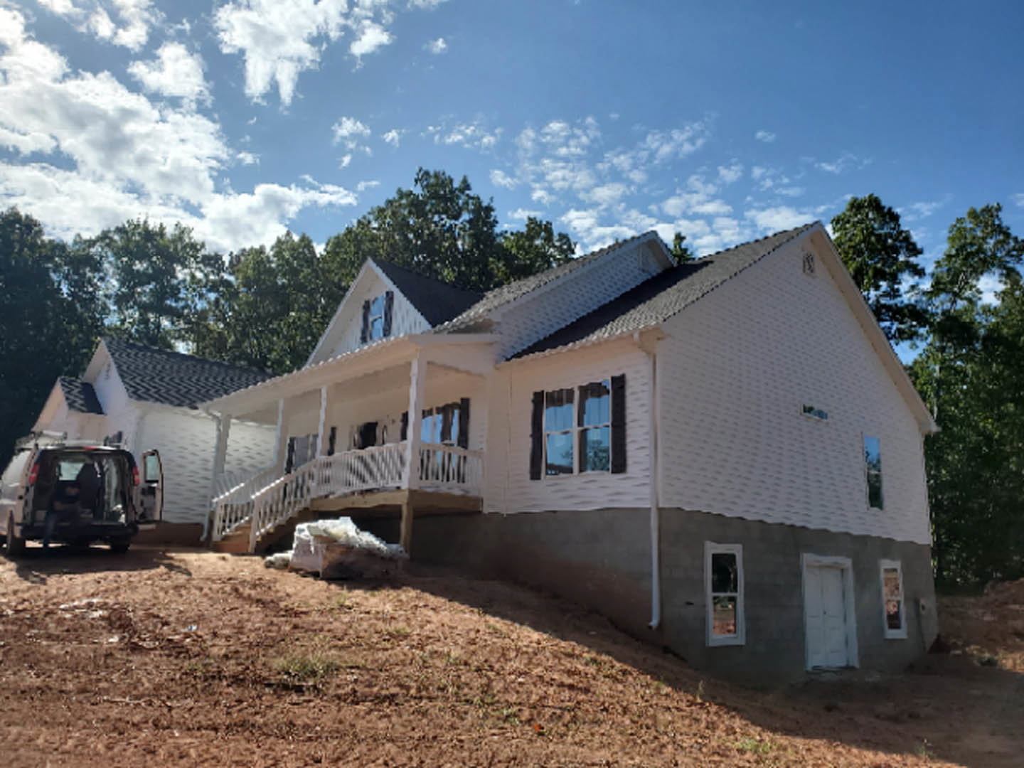 Partially built house with exposed framing, construction materials scattered on dirt ground, tractor parked nearby, large windows reflecting surrounding trees, cloudy sky overhead