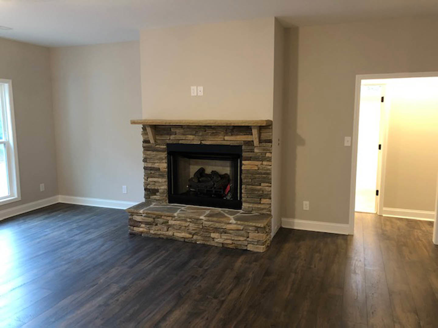 Modern living room with wood flooring, black glass fireplace set in white wall, wood mantel shelf, and sunlight streaming through nearby door and window