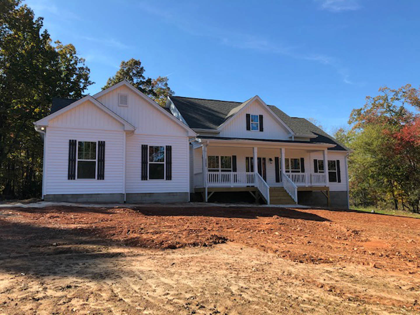 White house under construction with dirt foreground, trees in background, porch featuring white railing, multiple windows with white frames and black shutters