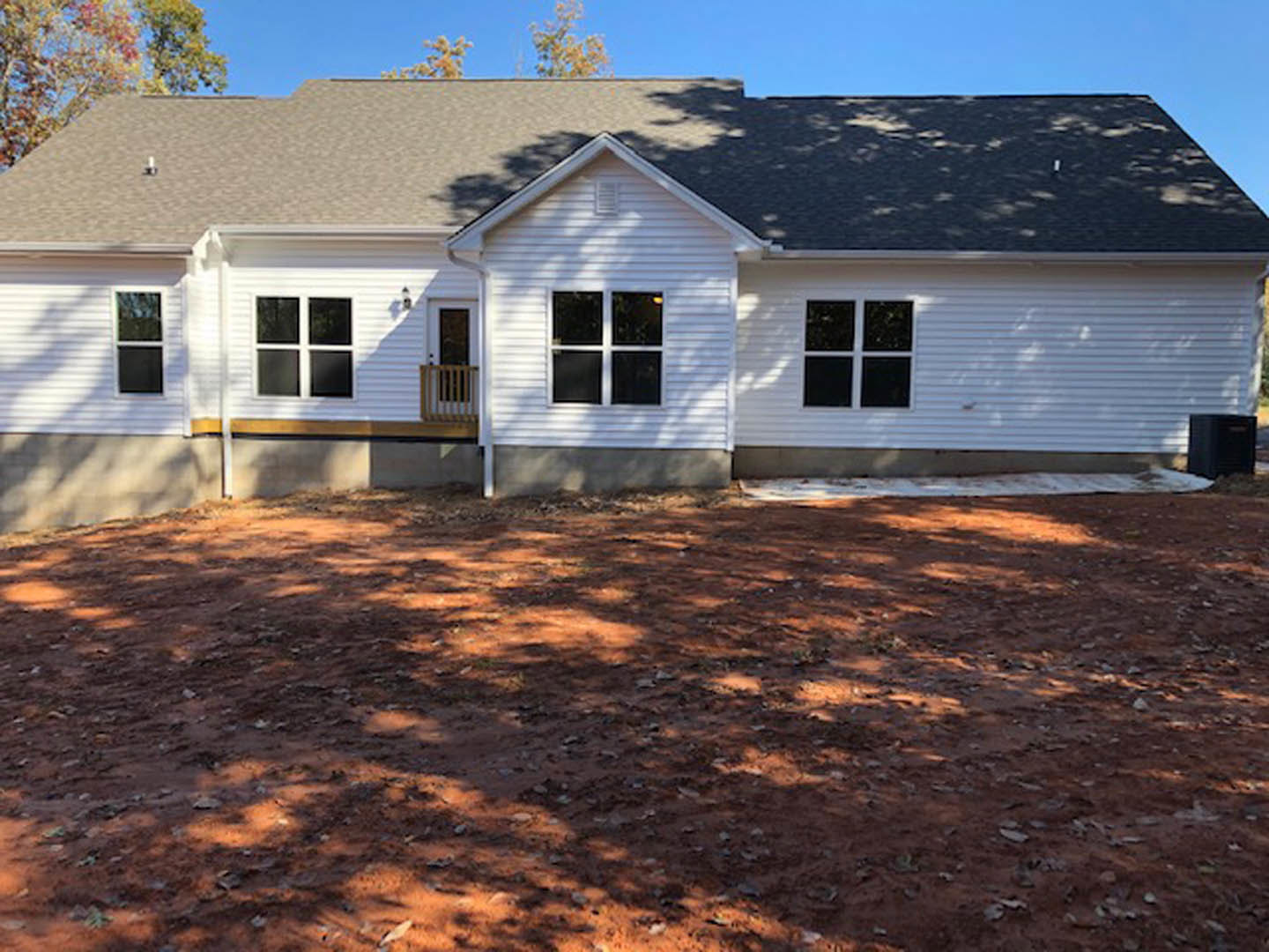White siding house with covered porch, large windows, and dirt yard scattered with leaves; blue sky and clouds overhead.