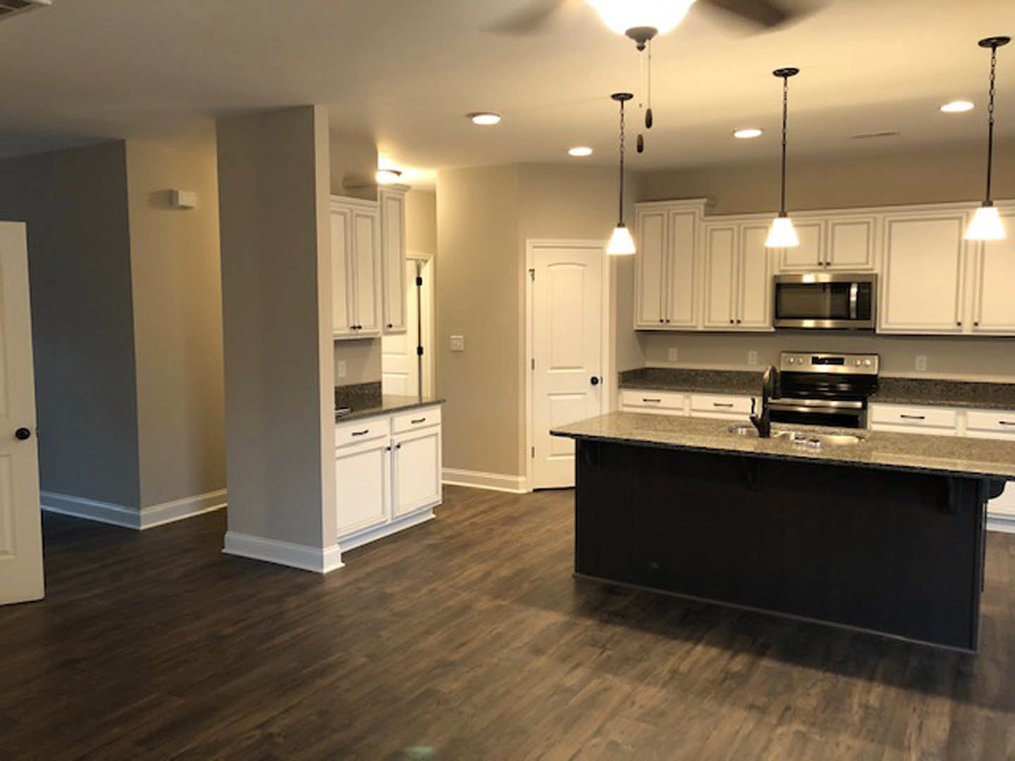 Spacious kitchen featuring a large central island with stone countertop, tile flooring, white cabinetry, stainless steel sink, and modern stove; white door with black hardware