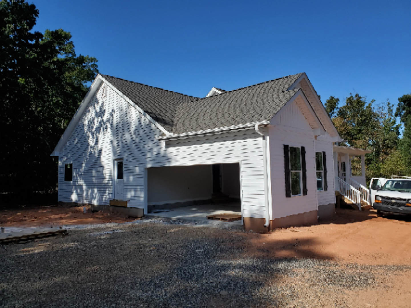 Framed house under construction with attached garage, white van parked in driveway, white-framed window, unfinished wall, black exterior light fixture, person standing on dirt lot