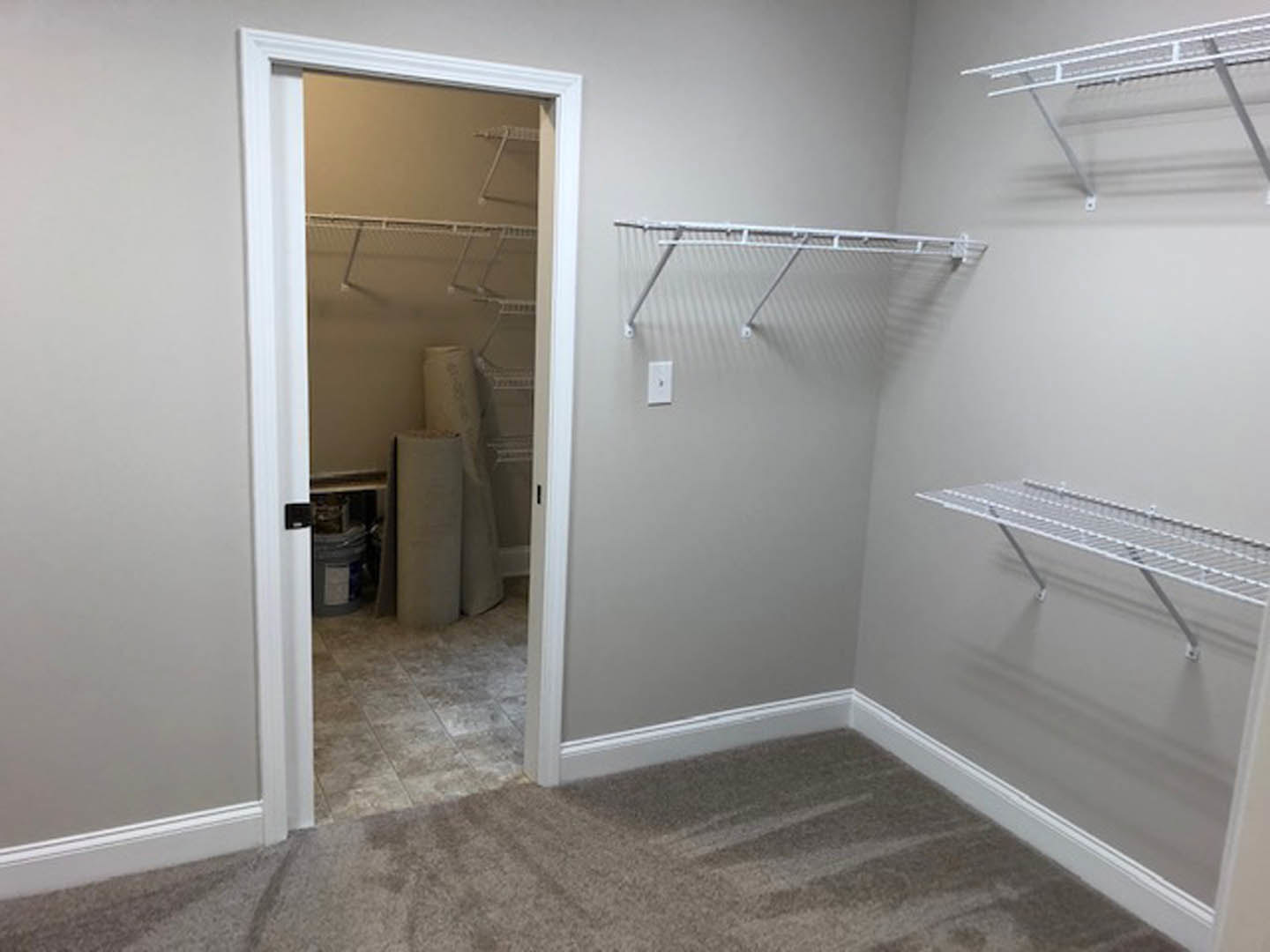Open closet with white metal shelves, hanging rod, and rolled-up paper stored on shelf; white plaster walls and door visible.