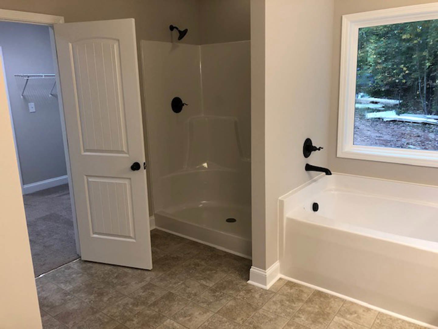 White bathroom featuring a freestanding bathtub with black faucet, glass-enclosed shower, window overlooking trees, white door with black knob, and black wall-mounted fixture on a