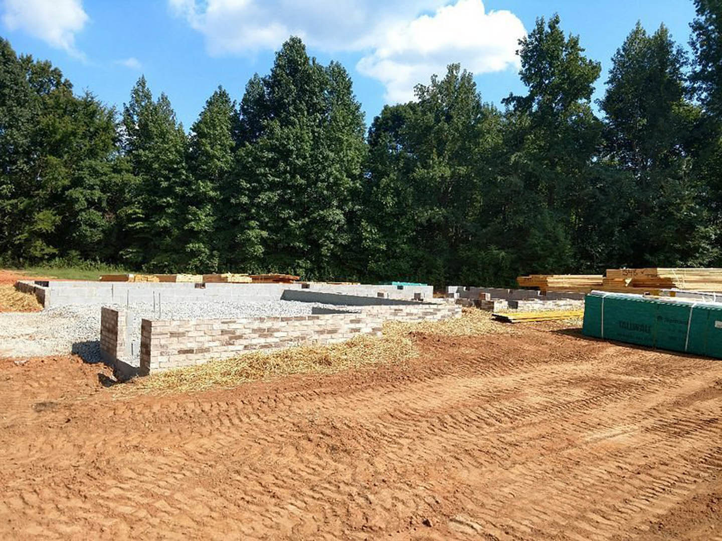 Partially built house framed with exposed wood, surrounded by dirt and sand, trees and blue sky with clouds in the background