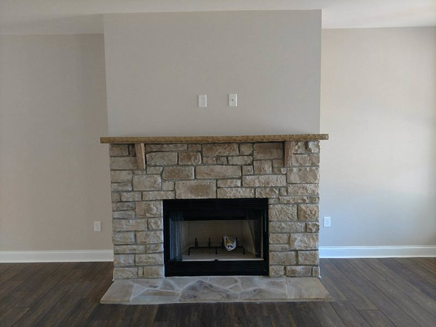 Stone fireplace with wood mantel shelf, newspaper resting on shelf, fire screen in front, stone hearth and floor, neutral wall in cozy living room.