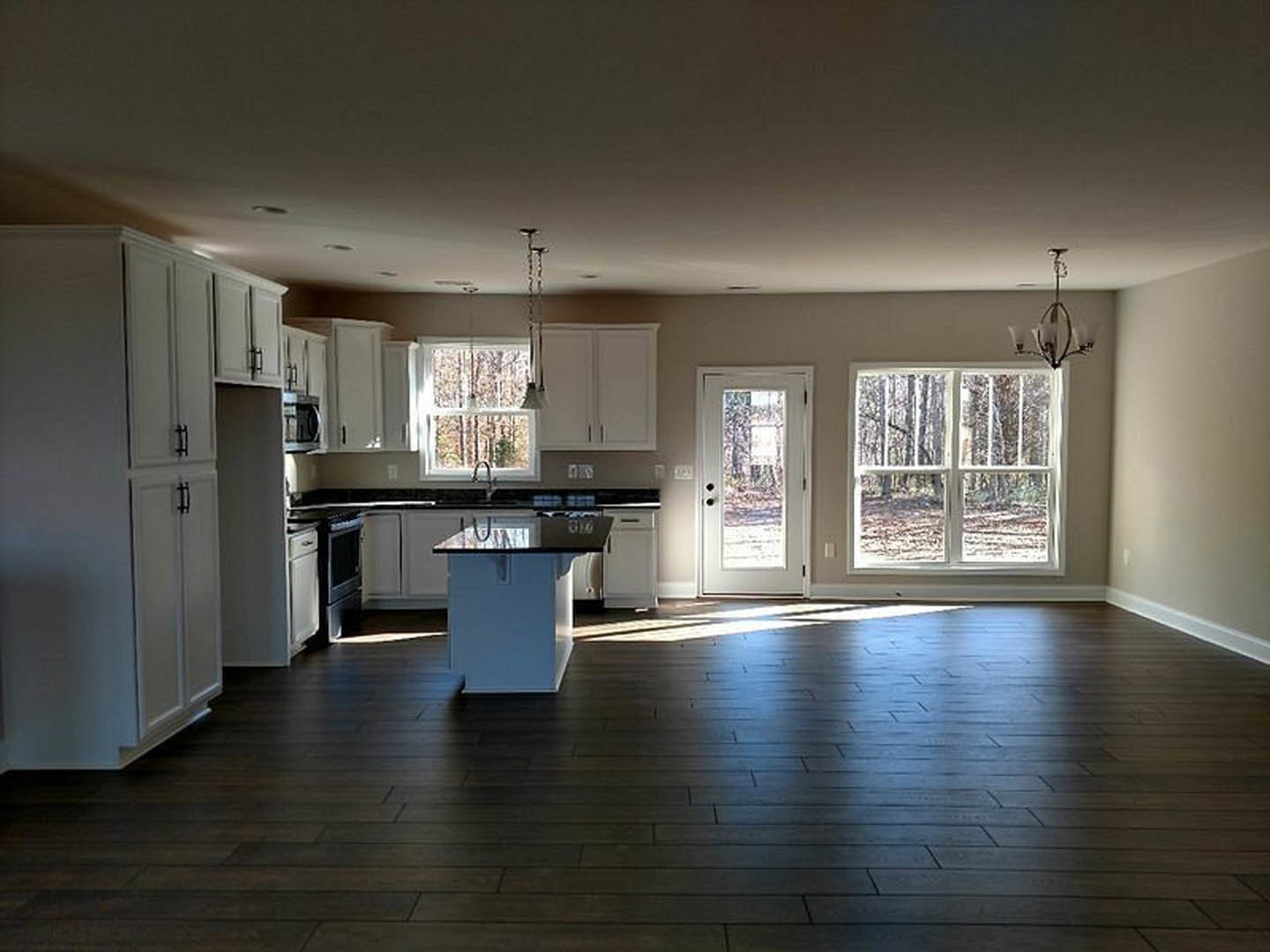 Large kitchen with dark wood flooring, central island featuring a black countertop, white cabinetry with black handles, white door with glass window, and window overlooking a