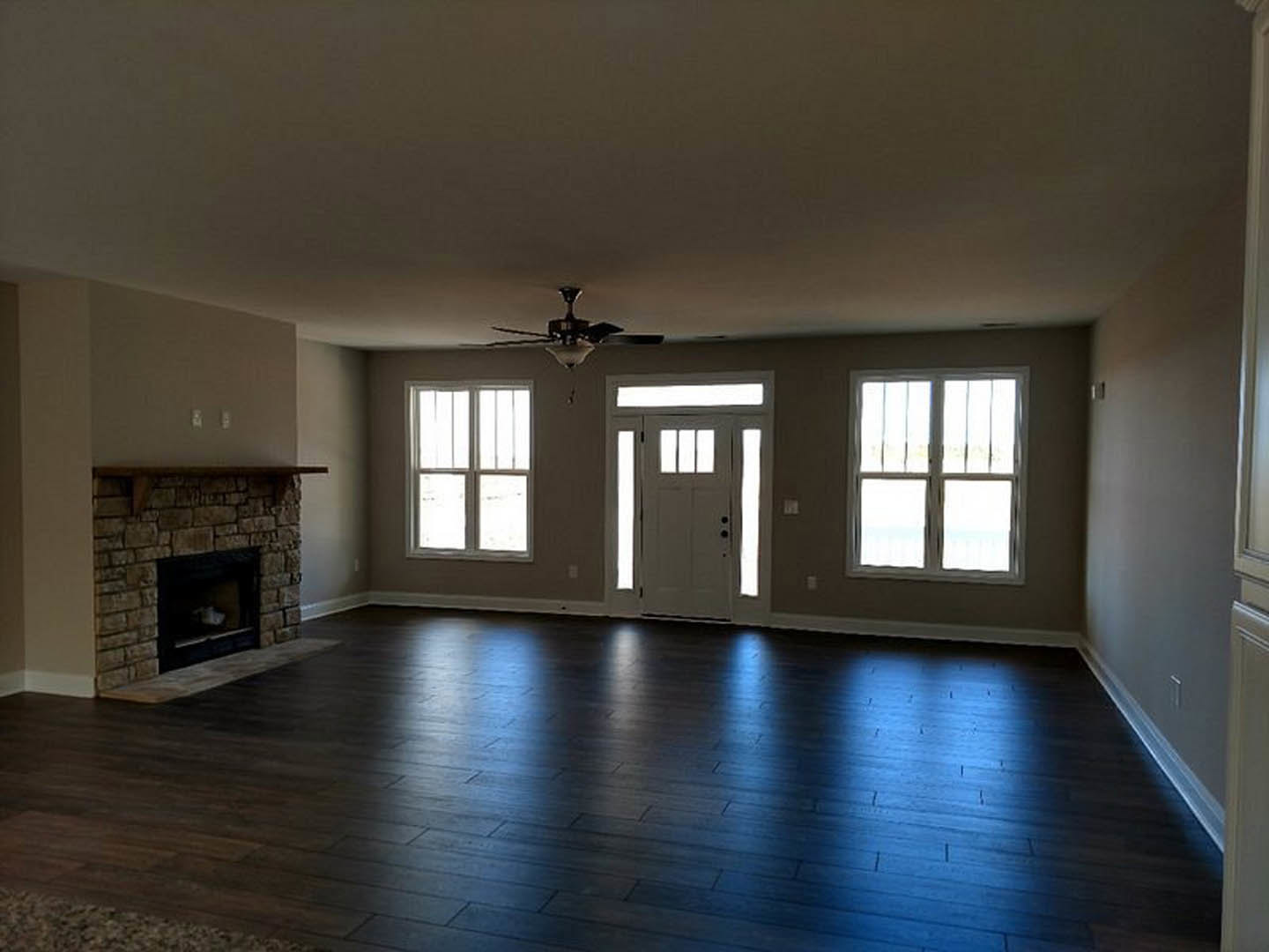 Living room with dark hardwood floors, stone fireplace, ceiling fan, white door with glass panes, and window with decorative bars