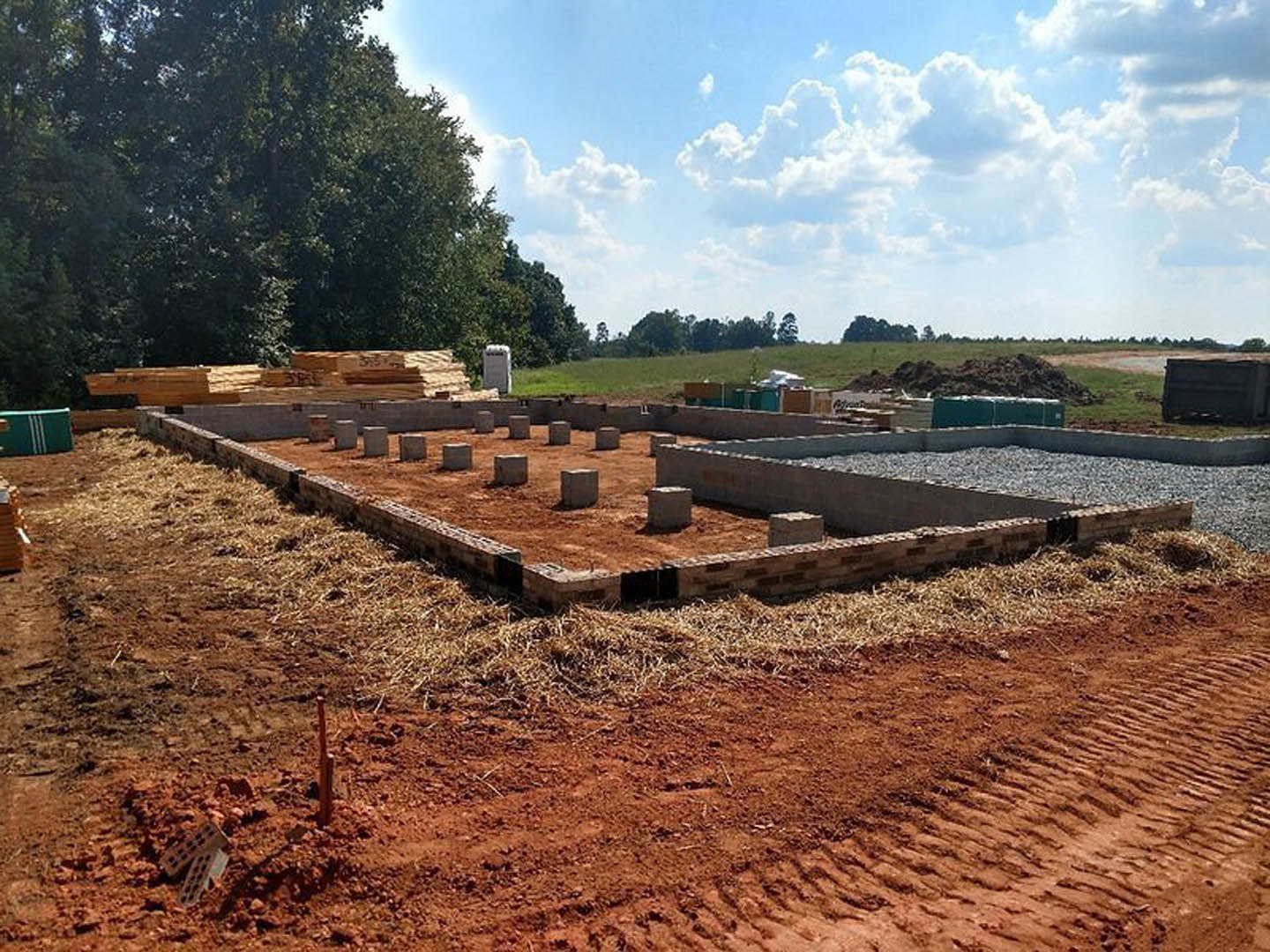 Construction site with exposed soil, scattered bricks, cement blocks, and surrounding trees under a partly cloudy blue sky