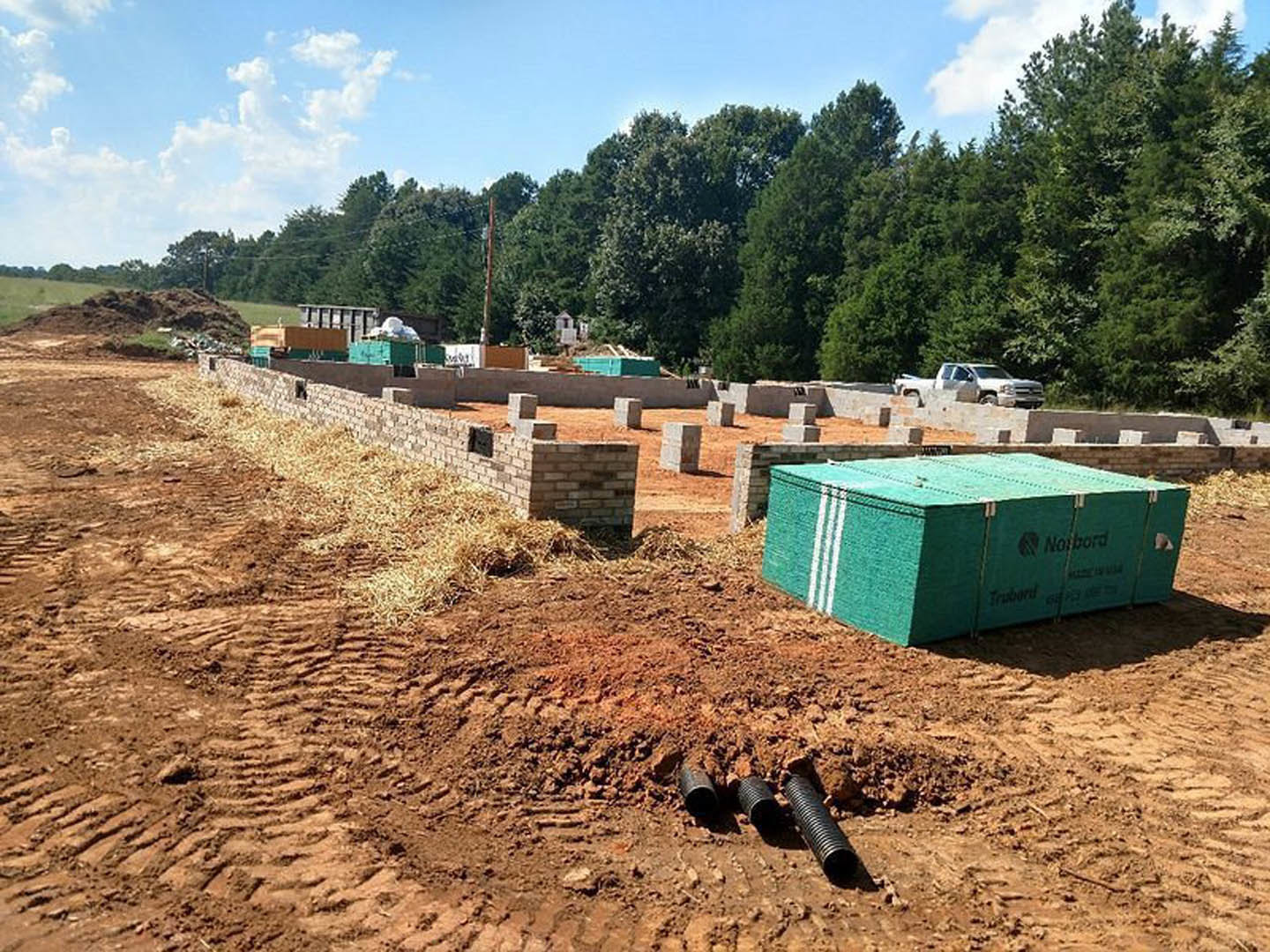 Concrete foundation slab with exposed plumbing pipes, green utility box, pile of dirt, white truck, and trees in the background under a partly cloudy sky