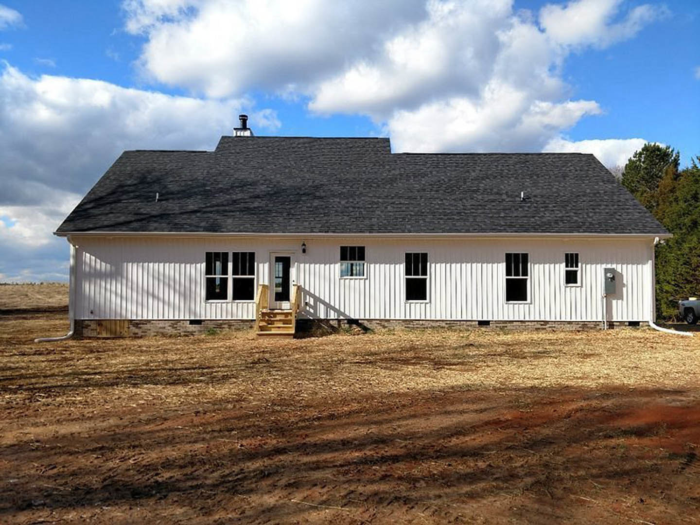 White farmhouse with black roof, white-framed windows, wooden staircase leading to dirt field, grassy lot under blue sky with scattered clouds, Wyckoff House visible in background.