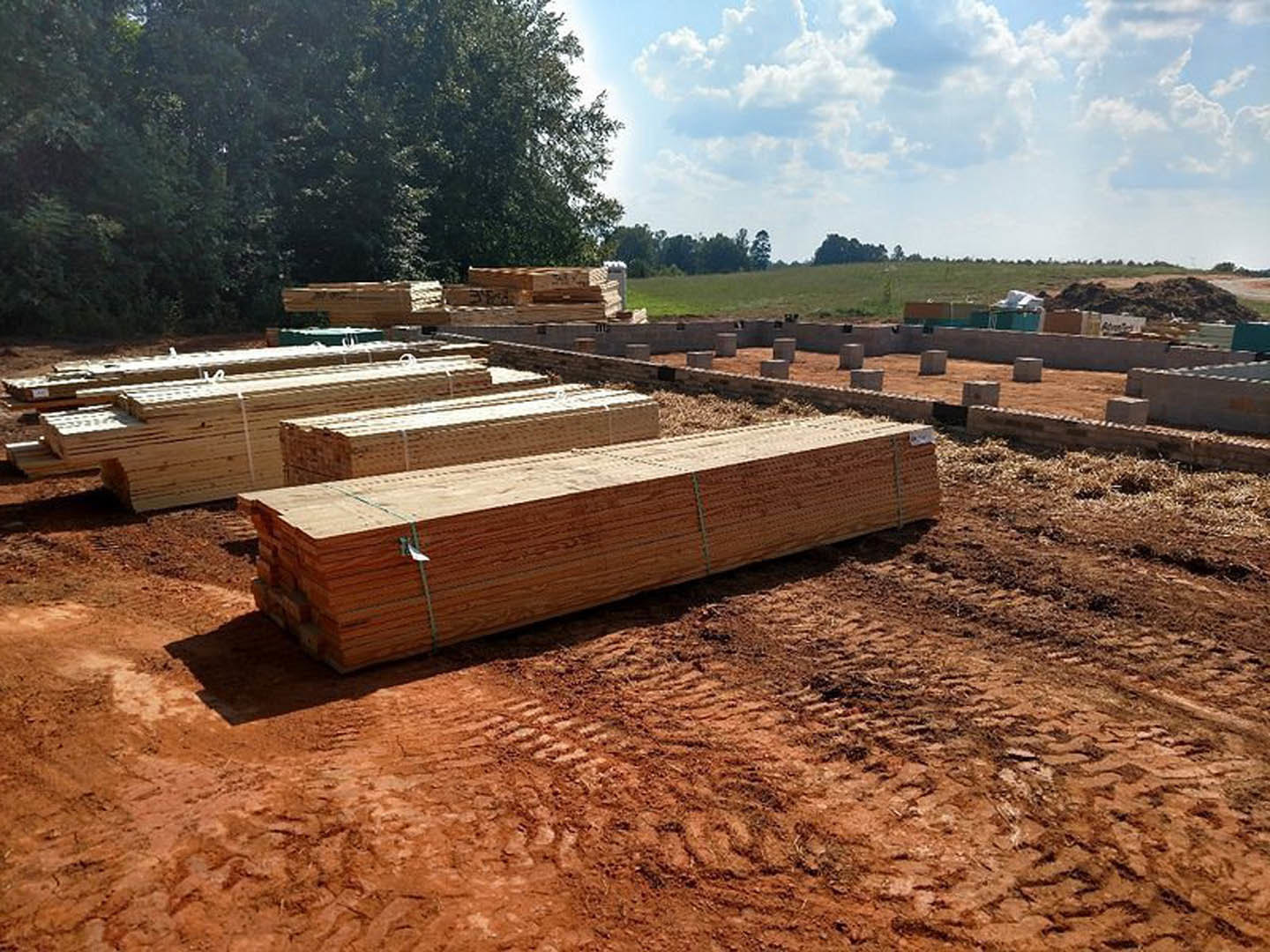 Stack of rough lumber on a dirt field under a partly cloudy blue sky, bordered by trees in the background