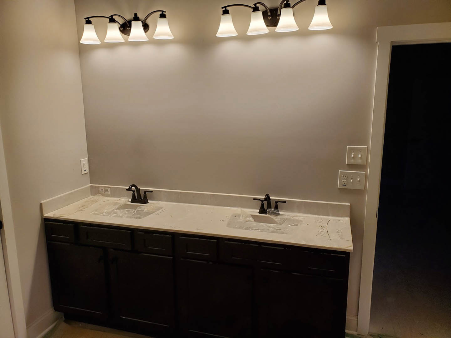 Bathroom with double white sinks, black hardware, wood vanity, large mirror, wall-mounted light fixture, and a white door with black handle