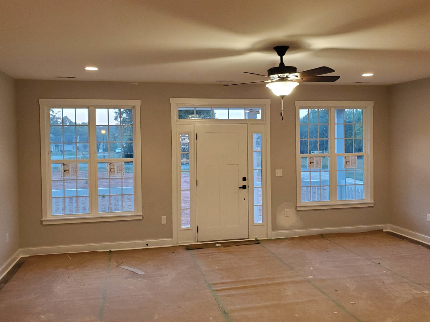 Spacious room with light-colored walls, ceiling fan, multiple large windows, white door with glass panes, and unfinished flooring with a roll of paper