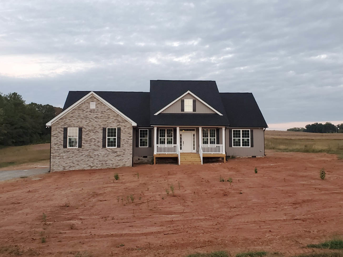 Brick house with black shutters, black roof, white door, and white railing, surrounded by dirt field and trees under cloudy sky