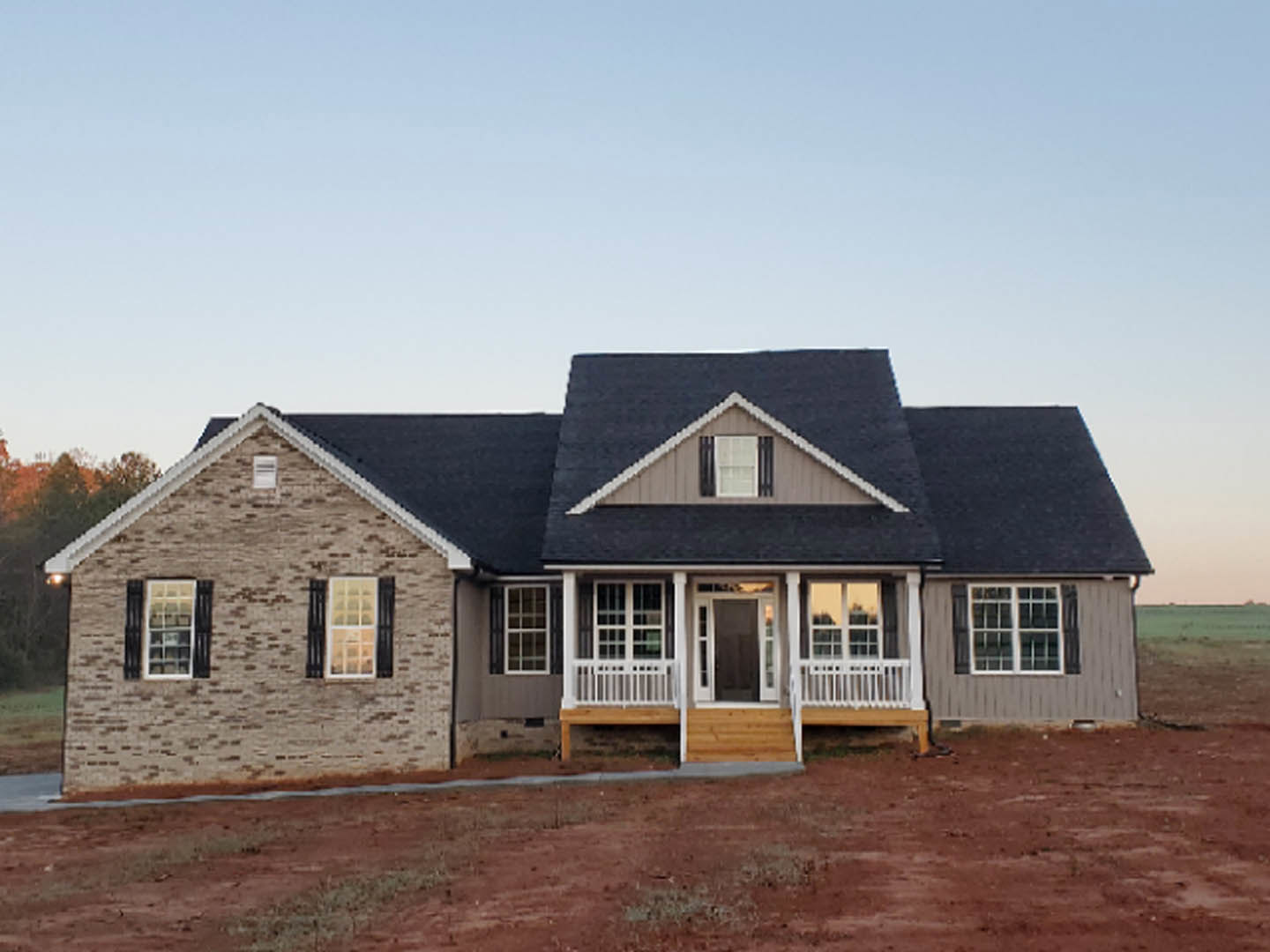 Brick house with black shutters and white window frames, covered porch, surrounded by red dirt yard under clear sky