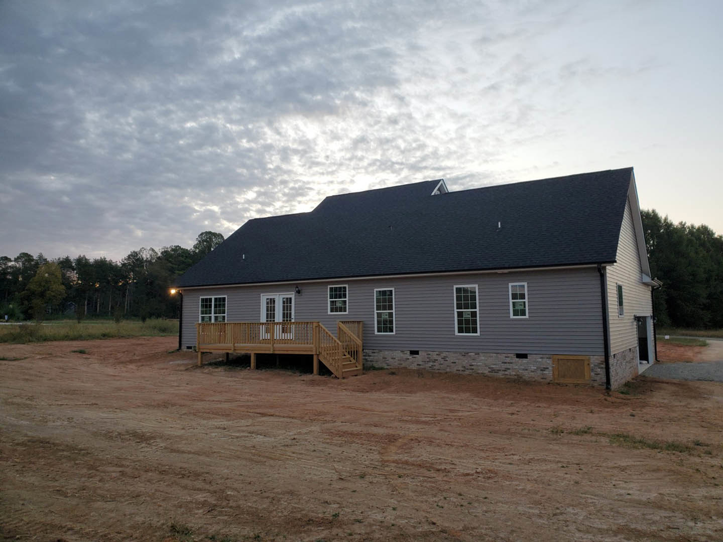 Two-story house with wood deck and stairs, surrounded by dirt road and open land, under a cloudy sky; multi-pane windows and railing visible.