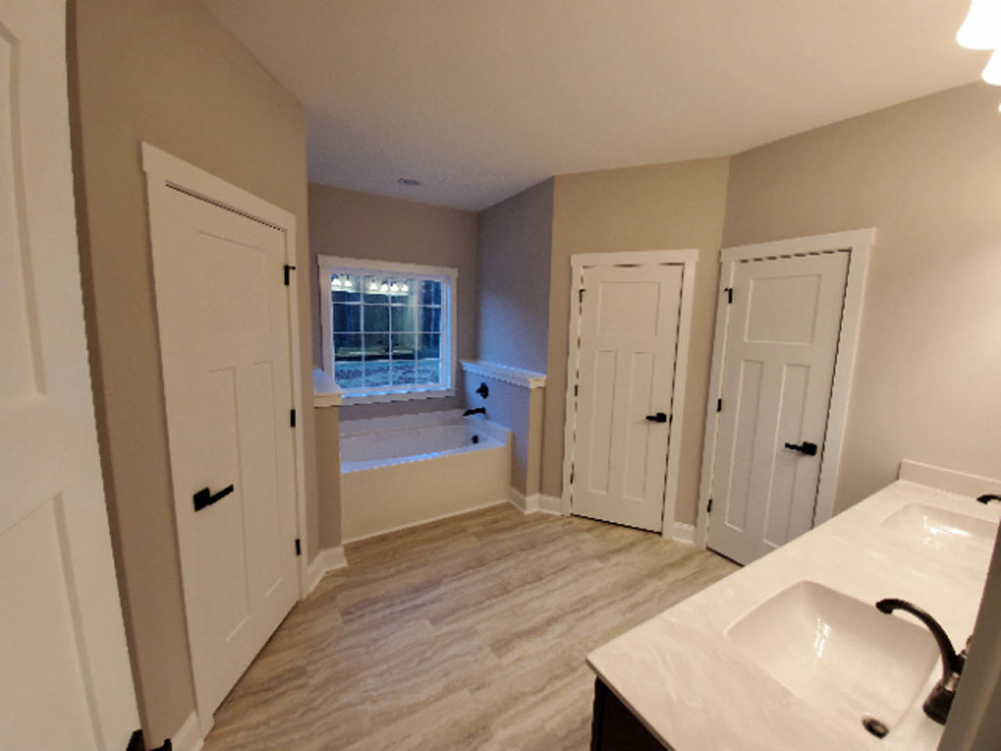 Bathroom featuring white paneled doors with black handles, freestanding soaking tub, white vanity with integrated sink, and large windows framed by white trim.