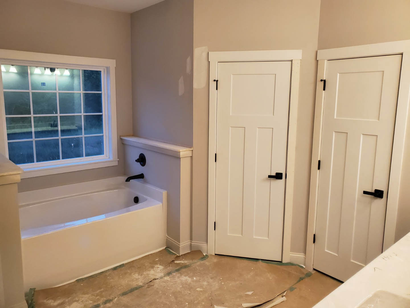 Bathroom with white paneled doors featuring black handles, white freestanding bathtub with matte black fixtures, light tile flooring showing a visible crack, and a row of windows